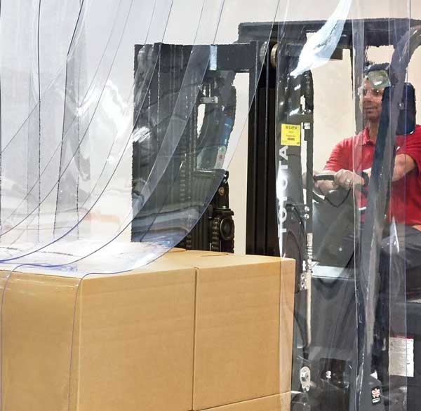 A forklift carrying a pallet loaded with boxes drives through a clear vinyl strip curtain covering a doorway.